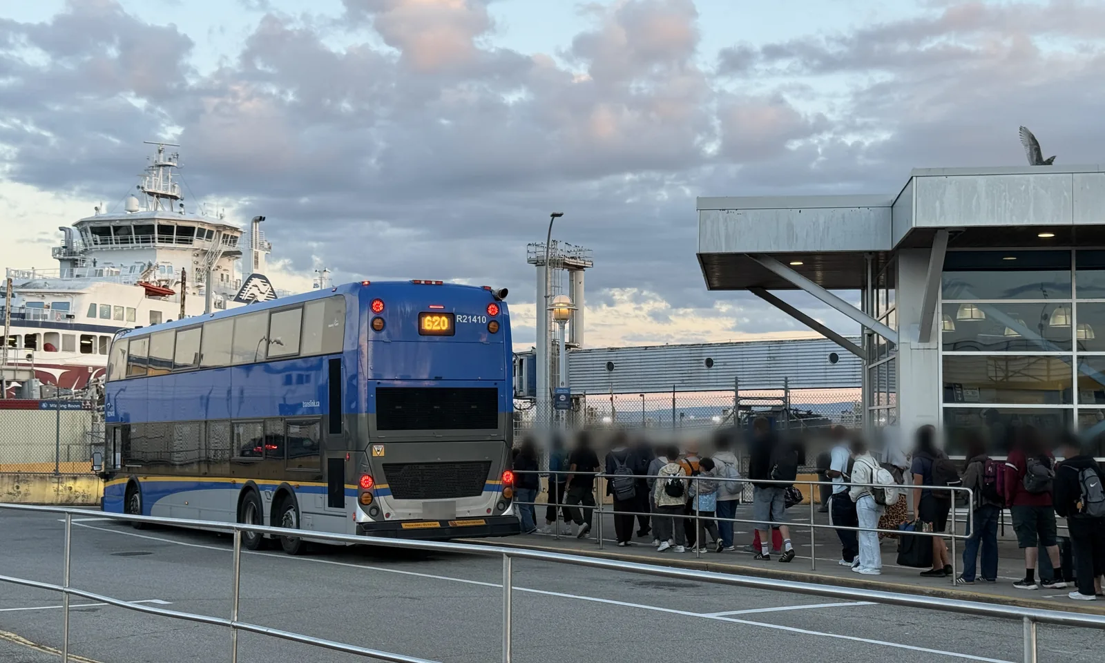 A double-decker 620 bus at the Tsawwassen Ferry Terminal bus stop, with passengers walking towards the terminal building and a ferry docked behind
